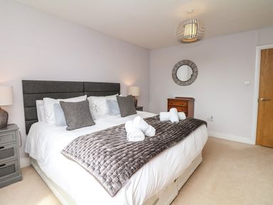 A bedroom with a double bed with grey cushions and a blanket a wooden chest of drawers a round mirror and two bedside tables with lamps at Nairn in Braunton