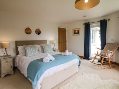 A bedroom with a bed with pillows and a blue throw blanket two bedside tables with lamps a wooden rocking chair near a window with dark curtains and a door at Nairn in Braunton