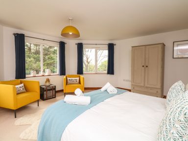 A bedroom with a bed covered with white and blue linens two yellow armchairs next to windows and a wooden wardrobe at Nairn in Braunton