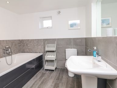A bathroom with a bathtub, toilet, sink, window, mirror, and a white basket shelf at Nairn in Braunton