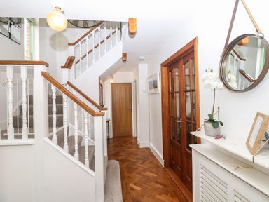 A hallway with wooden parquet flooring a staircase with white balusters and wooden handrails wooden doors a round mirror and a cabinet with a plant and picture frame at Nairn in Braunton