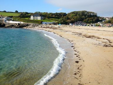 A sandy beach with small waves meeting the shore and houses on green hills at Hazel Barn in Falmouth