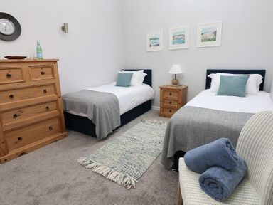 A bedroom with two single beds a wooden chest of drawers a wooden nightstand a chair with towels and three framed pictures on the wall at Hazel Barn in Falmouth