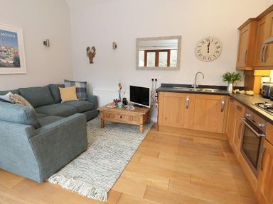 A living area with blue sofa a wooden coffee table a small television a wall mirror and a kitchen with wooden cabinets at Hazel Barn in Falmouth