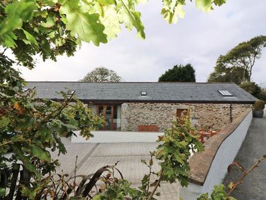 A stone barn with slate roof and patio with outdoor furniture seen through green leaves at Hazel Barn in Falmouth