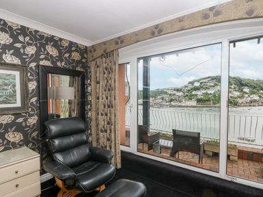 A room with black leather chair and ottoman a dresser with drawers floral wallpaper and a balcony with chairs overlooking a water body at Bight Boathouse in Dartmouth