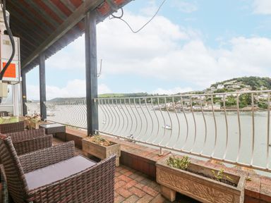 A balcony with wicker chairs and planters overlooking a river and houses on a hillside at Bight Boathouse in Dartmouth