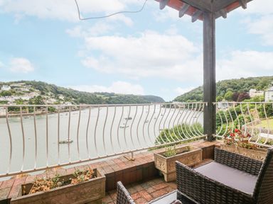 A balcony with two wicker chairs and planters overlooking a river with boats and houses on hills at Bight Boathouse in Dartmouth