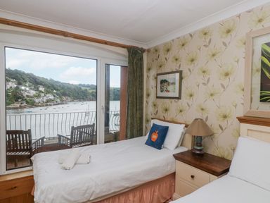 A bedroom with two single beds a nightstand and a balcony overlooking a river at Bight Boathouse Dartmouth