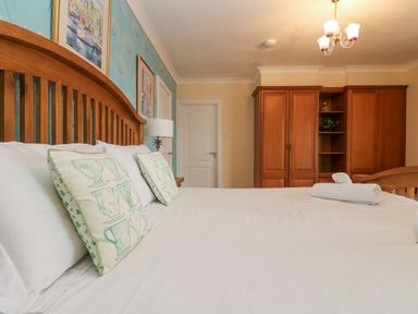 A bedroom with a wooden bed frame white bed linen pillows with teacup designs blue floral wallpaper wooden wardrobe and ceiling light at Bight Boathouse in Dartmouth