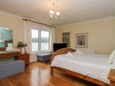 A bedroom with a wooden bed and white bedding a wooden dressing table with a mirror and flowers and a window showing a view of boats on water at Bight Boathouse in Dartmouth