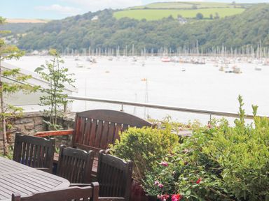 A wooden outdoor dining table with chairs and a bench on a terrace overlooking a body of water with boats and hills in the background at Bight Boathouse Dartmouth
