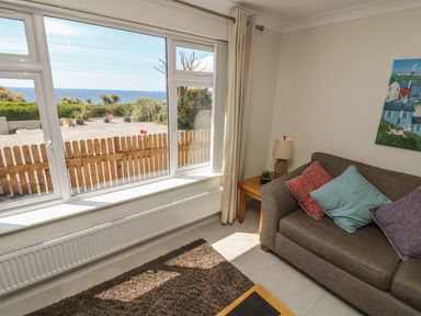 A living room with a brown sofa with cushions a side table with a lamp a large window showing a garden and sea view at Hagar Vor in Marazion
