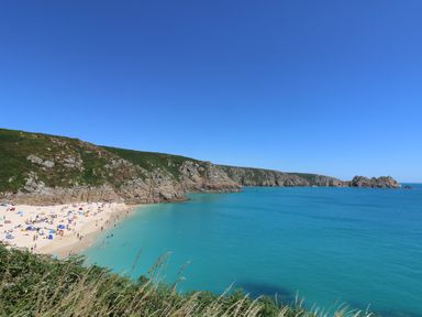A beach with people on sand and swimming in the water by rocky cliffs and clear sky at Hagar Vor in Marazion