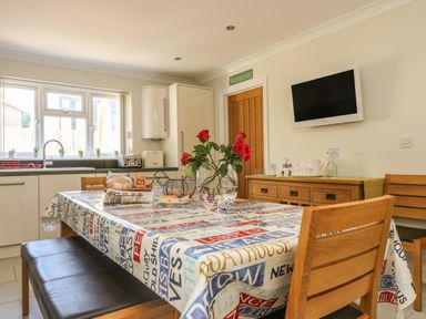 A kitchen dining area with a table covered by a printed tablecloth with chairs bench a vase with red flowers a sideboard with tea set a wall mounted tv and a washroom door at Hagar Vor in Marazion