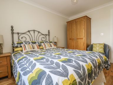 A bedroom with a bed featuring patterned bedding an iron headboard wooden bedside tables with lamps a wooden wardrobe and a chair with a cushion at Hagar Vor in Marazion