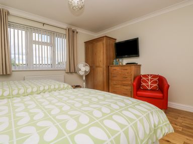 A bedroom with two beds with patterned bedspreads a wooden wardrobe a wooden dresser with a wall-mounted TV a red armchair and a window with curtains and blinds at Hagar Vor in Marazion