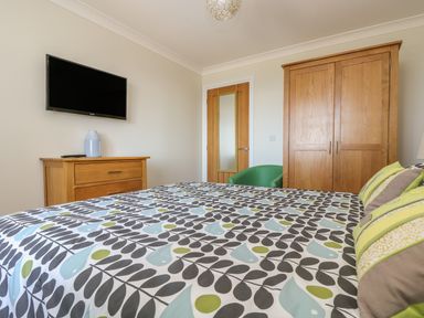 A bedroom with a bed with patterned bedding a wooden dresser with a vase a wall-mounted TV a wooden wardrobe a green chair and a door with a mirror panel at Hagar Vor in Marazion