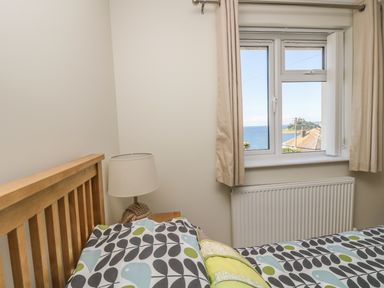A bedroom with a wooden bed frame a lamp on a nightstand and a window with curtains showing a view of the sea at Hagar Vor in Marazion