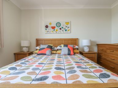 A bedroom with a wooden bed and colorful leaf-patterned bedding flanked by two wooden nightstands with lamps and a wooden dresser at Hagar Vor in Marazion