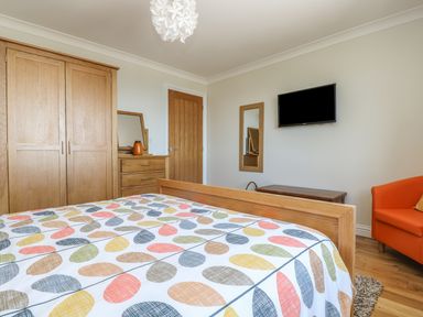 A bedroom with a bed covered in a patterned bedspread a wooden wardrobe wooden chest of drawers a wall-mounted television an orange armchair and a wooden door at Hagar Vor in Marazion