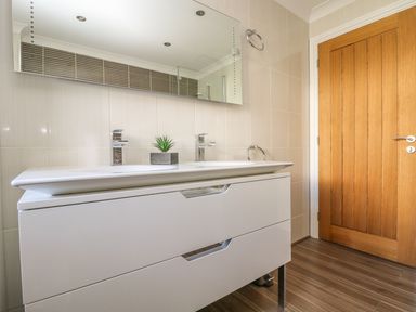 A bathroom with a white double sink vanity a rectangular mirror a small plant on the counter a wooden door and towel rings on tiled walls at Hagar Vor in Marazion