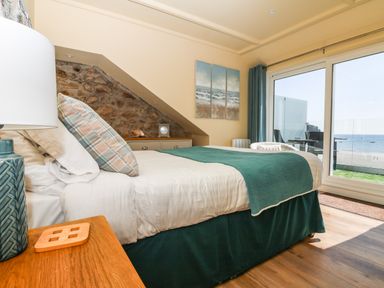 A bedroom with a bed covered by a teal blanket and pillows a wooden nightstand with a lamp a stone wall an ocean view through sliding glass doors at Higher Mount View in Marazion