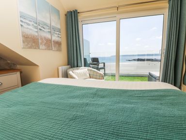 A bedroom with a bed covered in a green quilt and a sliding glass door opening to a balcony with chairs overlooking a beach at Higher Mount View in Marazion