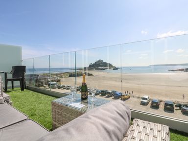 A balcony with seating and a table with a bottle and glasses overlooking a beach and an island at Higher Mount View in Marazion