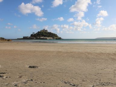 A sandy beach with a small island featuring buildings and trees in the sea under a partly cloudy sky at Higher Mount View in Marazion
