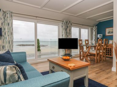 A living room with a blue sofa and wooden coffee table next to large windows overlooking the beach at Higher Mount View in Marazion