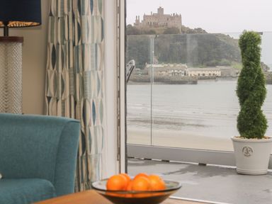 A room with a blue chair a curtain a table with a bowl of oranges and a view of a castle and water at Higher Mount View in Marazion