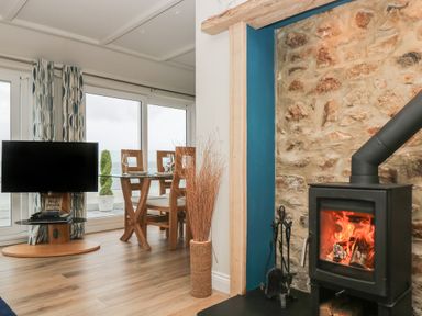 A living area with a wood stove fireplace a glass dining table with chairs a television and large windows at Higher Mount View in Marazion