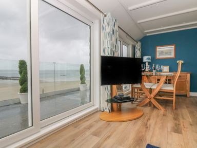 A dining area with a wooden table and chairs next to a television in front of large windows overlooking a beach at Higher Mount View in Marazion