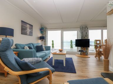A living room with blue chairs and sofa a wooden coffee table and a TV near large windows with a sea view at Higher Mount View in Marazion