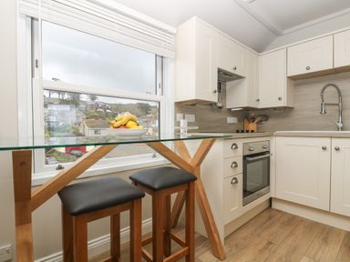 A kitchen with white cabinets a glass table with wooden legs two black stools and a bowl of fruit on Higher Mount View in Marazion