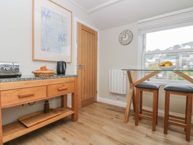 A kitchen with a wooden sideboard holding a toaster croissants and a kettle a glass table with fruit bowl and two stools at Higher Mount View in Marazion