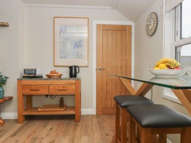 A kitchen area with a wooden table holding a toaster croissants and a kettle a wooden door two stools and a glass table with a fruit bowl at Higher Mount View in Marazion
