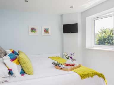 A bedroom with a white bed with colorful pillows a wooden tray with strawberries flowers and magazines and a wall-mounted tv by a window at Lake View in Kingsand and Cawsand