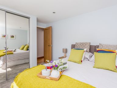A bedroom with a bed, pillows, a yellow throw blanket, a wooden tray with bowls and flowers, a wooden door, a bedside table with a lamp, and mirrored sliding closet doors at Lake View in Kingsand and Cawsand
