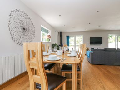A dining area with wooden table and chairs set with white dishes and wine glasses next to a living room with gray sofas and a wall mounted TV at Lake View in Kingsand and Cawsand