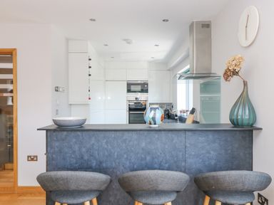 A kitchen with a dark countertop bar with three gray stools and kitchen appliances in the background at Lake View in Kingsand and Cawsand