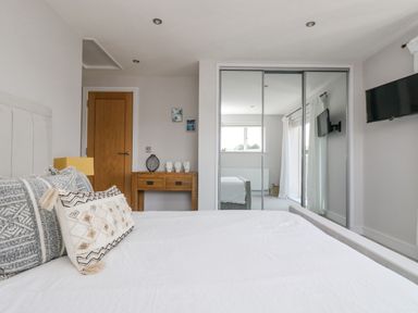 A bedroom with a bed and decorative pillows a wooden door a wooden dresser with decor items mirrored closet doors and a wall-mounted television at Lake View in Kingsand and Cawsand