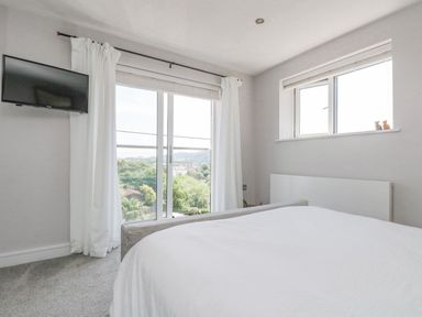 A bedroom with a bed white curtains a mounted TV and windows showing trees at Lake View in Kingsand and Cawsand