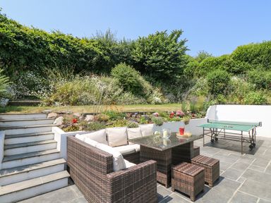 An outdoor patio with wicker seating and a table beside a ping pong table and stairs leading to a garden at Lake View Kingsand and Cawsand