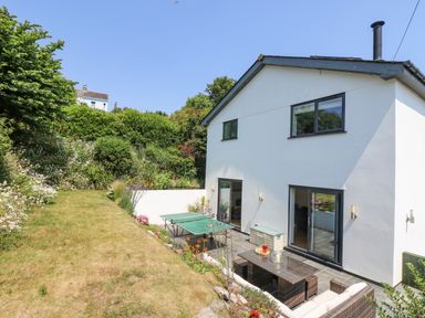 A garden with grass and flowers next to a white two-story house with a patio and outdoor furniture at Lake View in Kingsand and Cawsand
