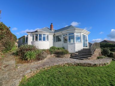 A white house with large windows stone steps and a stone path surrounded by grass and shrubs at Gwillen in Mawgan Porth