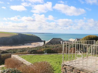A coastal garden area with a stone patio and metal railing overlooking houses cliffs and the sea at Gwillen in Mawgan Porth