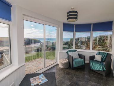 A sunroom with two armchairs and a side table by large windows overlooking a coastal landscape at Gwillen in Mawgan Porth