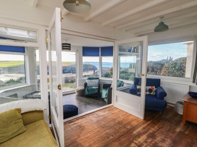 A living room with armchairs and a sofa opening to a sunroom with chairs and poufs with a view of the coast at Gwillen in Mawgan Porth